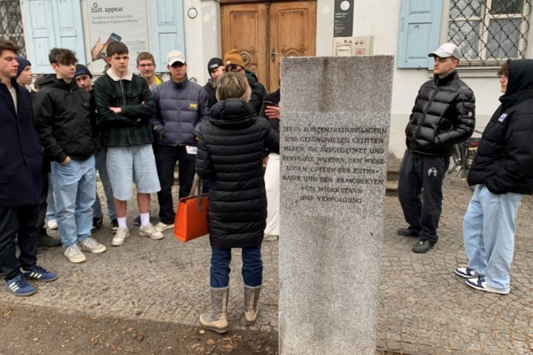 Eine Gruppe Jugendlicher steht um eine Frau, die vor einem Gedenkstein spricht. Die Inschrift erinnert an die Opfer von Konzentrationslagern, Euthanasie und politischer Verfolgung. Die Szene spielt sich vor einem historischen Gebäude mit blauen Fensterläden ab. Die Jugendlichen tragen Freizeitkleidung, einige hören aufmerksam zu, andere haben die Arme verschränkt oder die Hände in den Taschen. © Christoph Rinderer